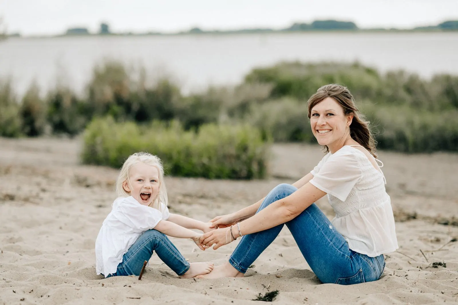 Mutter und Tochter sitzen barfuß im Sand an der Elbe und halten sich an den Händen