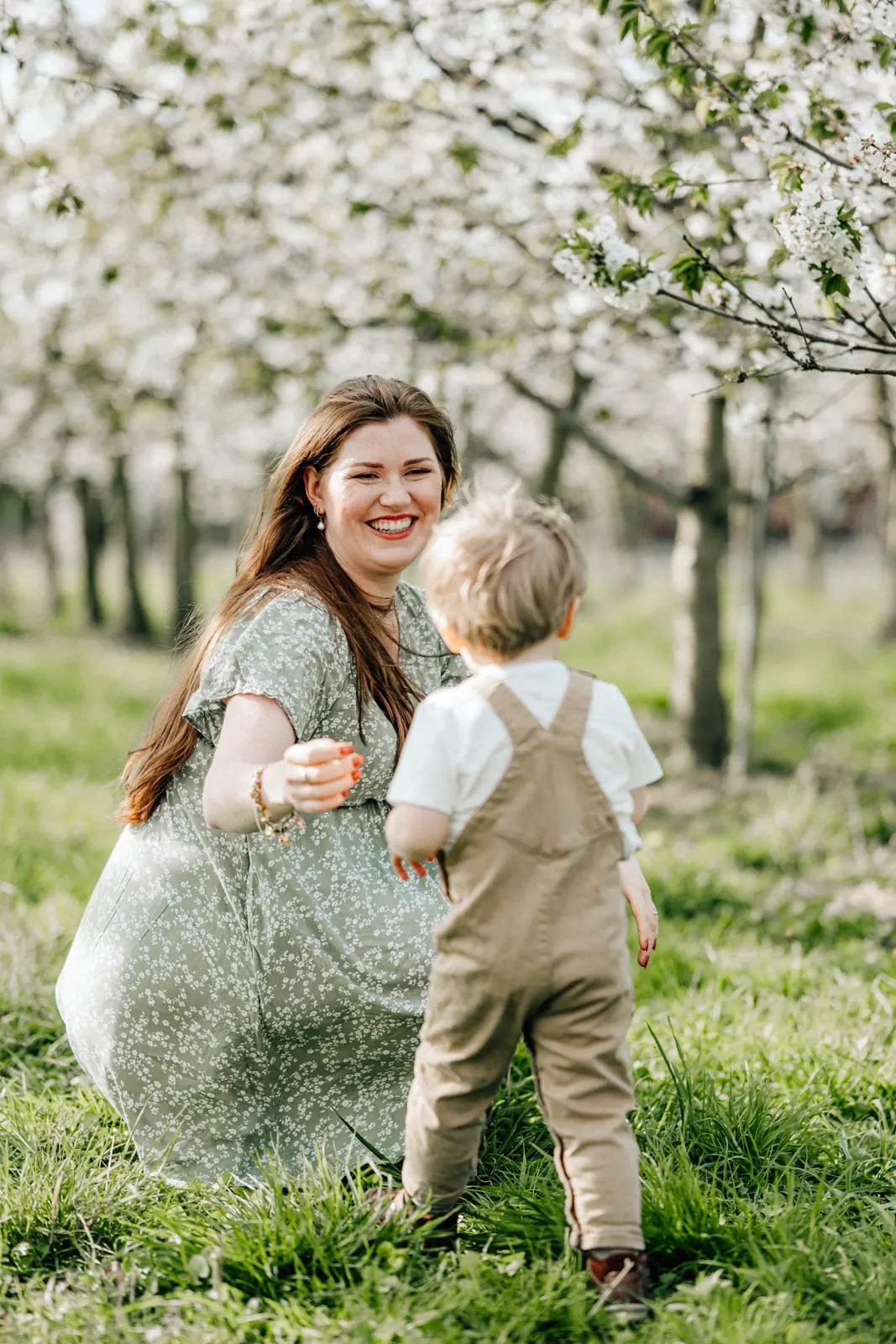 Mutter streckt die Arme nach ihrem kleinen Sohn aus, Obstblüte im Hintergrund