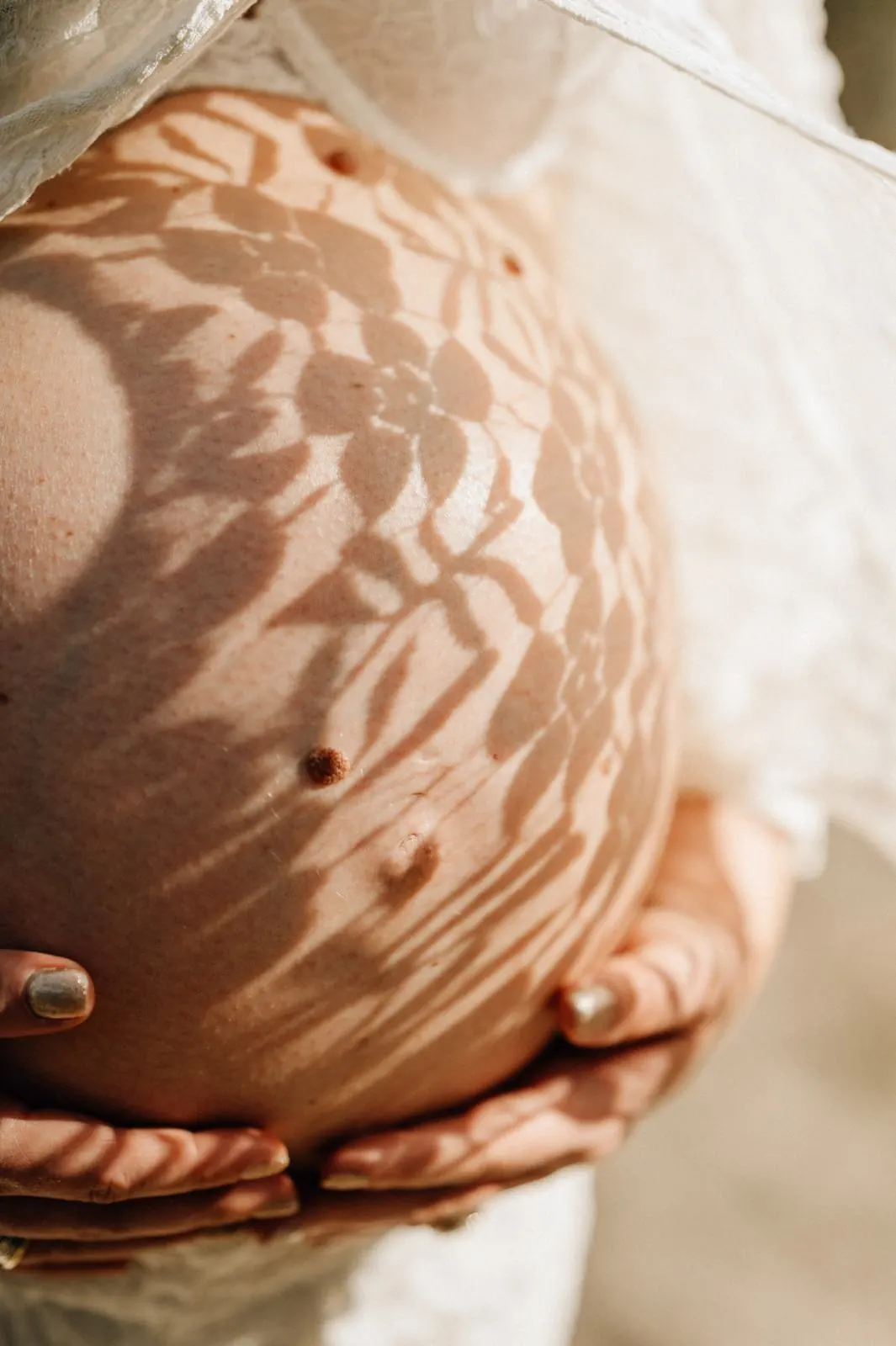 Babybauch mit Schattenmuster aus Spitze, zwei Hände halten den Bauch